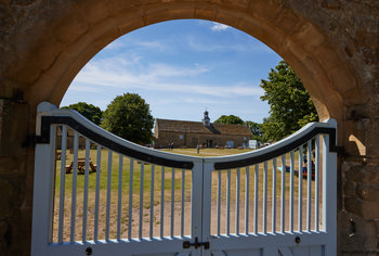 Hardwick Hall yard This architectural landscape photograph shows the yard of Hardwick Hall, a prominent Elizabethan manor house in Derbyshire, United Kingdom. Taken on a summer morning, the image is framed through a stone archway and light blue gate that opens onto a grassy yard with picnic tables and people enjoying the outdoors. In the background, the distinctive clock tower and stone-built stables associated with the National Trust site are visible, set amongst mature trees under a blue sky. Hardwick Hall, renowned for its Elizabethan architecture, is a notable historical landmark managed by the National Trust, reflecting the heritage of Derbyshire and the United Kingdom.